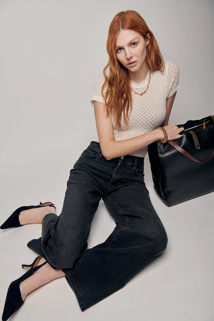 A woman lies on the floor, showcasing a mesh lace baby tee and relaxed-fit black denim from Frame. The cool, laid-back vibe is complemented by accessories from Jenny Bird, including a gold bracelet and necklace, adding an air of sophistication. A large black passenger bag from Rag & Bone completes the scene, creating an urban and effortlessly stylish aesthetic. The editorial setting captures the model's confidence, blending casual and refined elements in a unique and captivating composition.