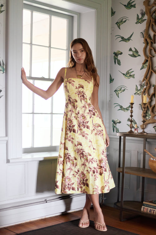 Woman in a floral dress standing in a room with decorative wallpaper and a window.