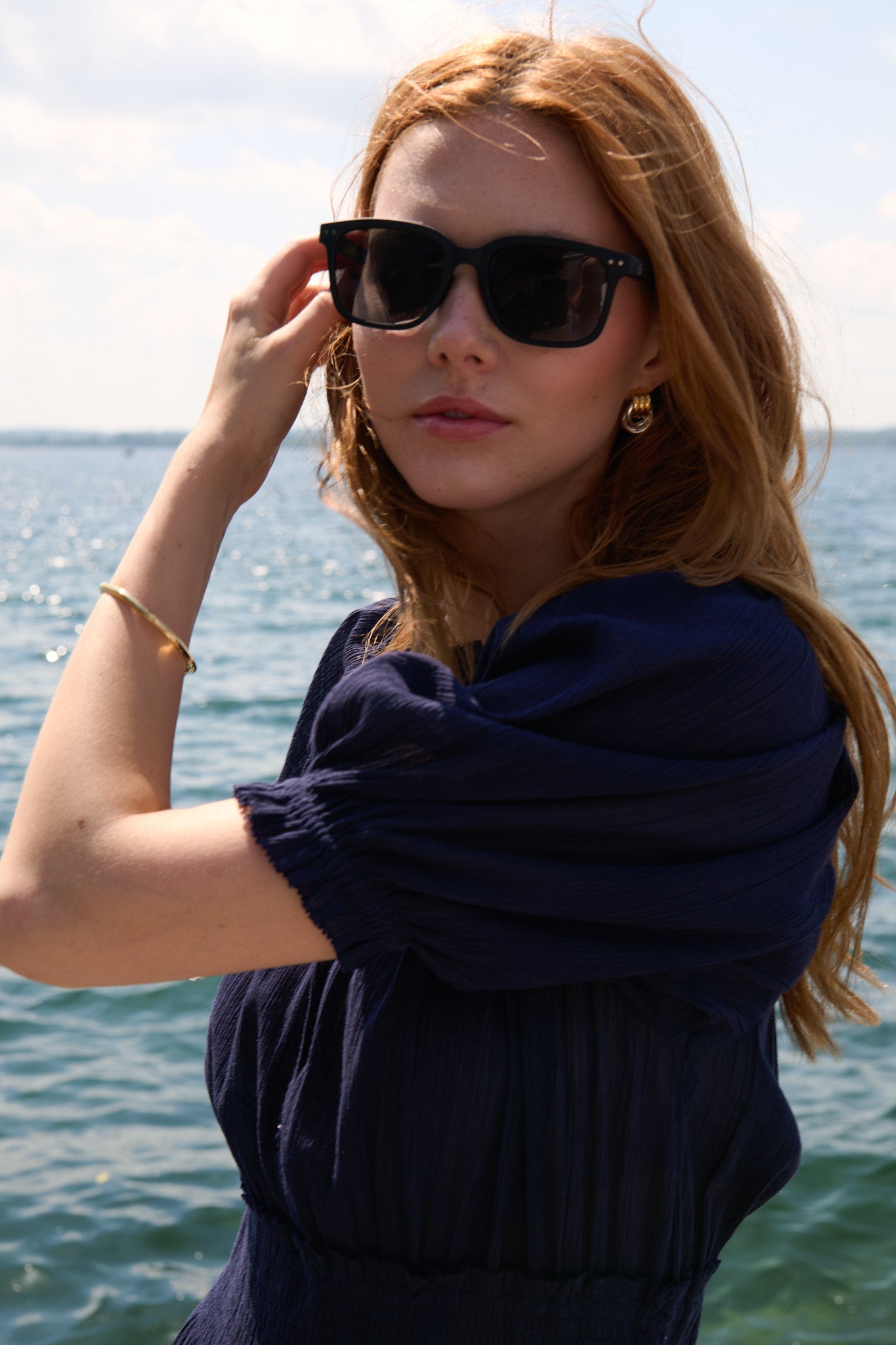 Close lakeside portrait of a model in a navy Apiece Apart dress with sunglasses and gold jewellery