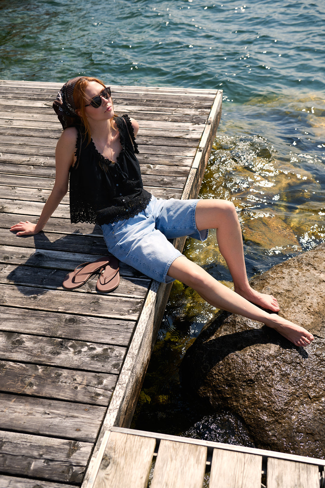 Model in a black DÔEN eyelet top and AG Jeans shorts seated barefoot beside the lake