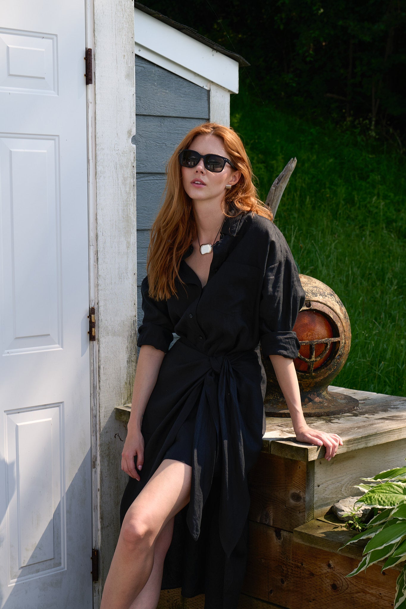 Model in a black Apiece Apart shirt dress leaning beside a grey lakeside boathouse
