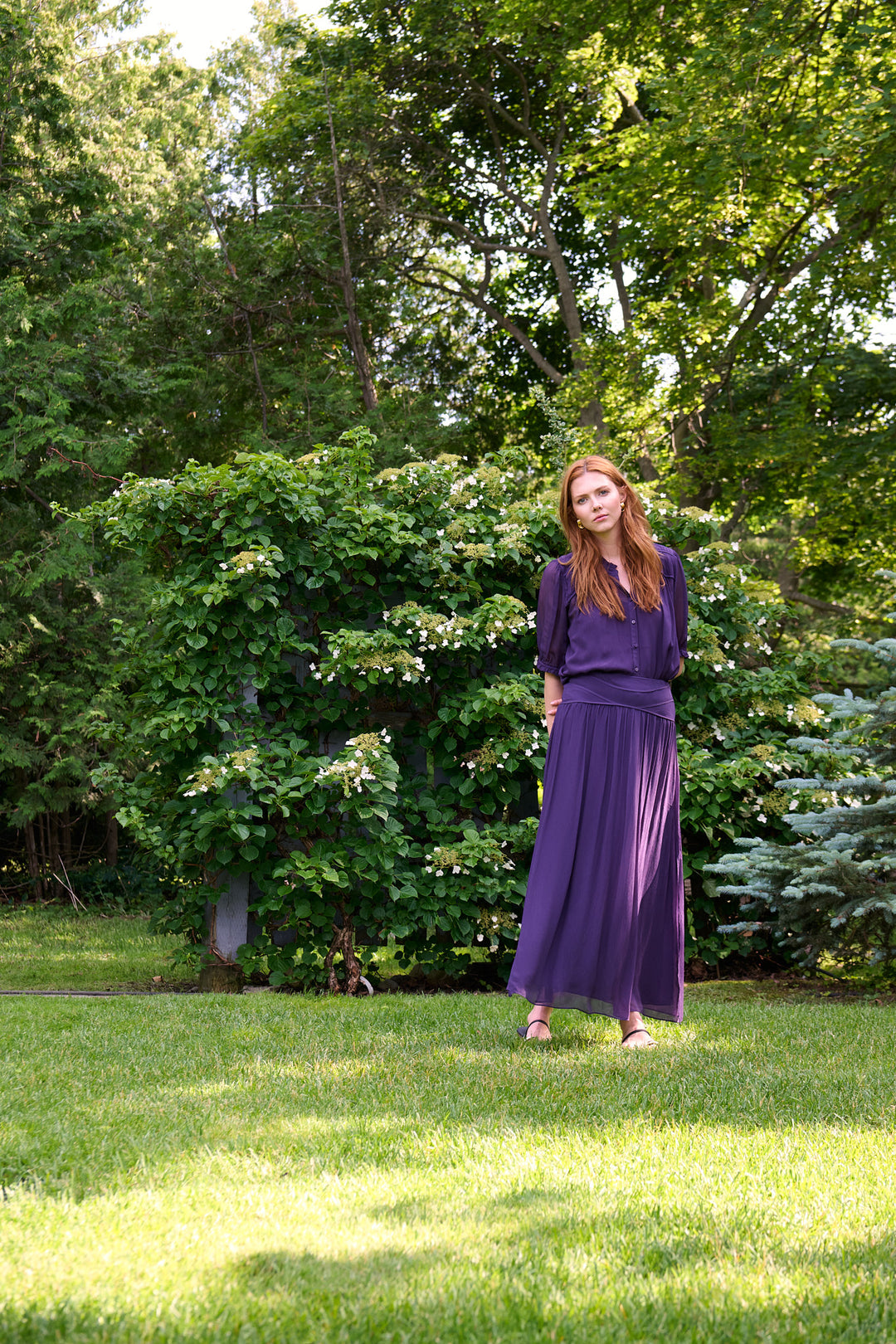 Wide garden portrait of a model in a tonal plum ba&sh blouse and maxi skirt