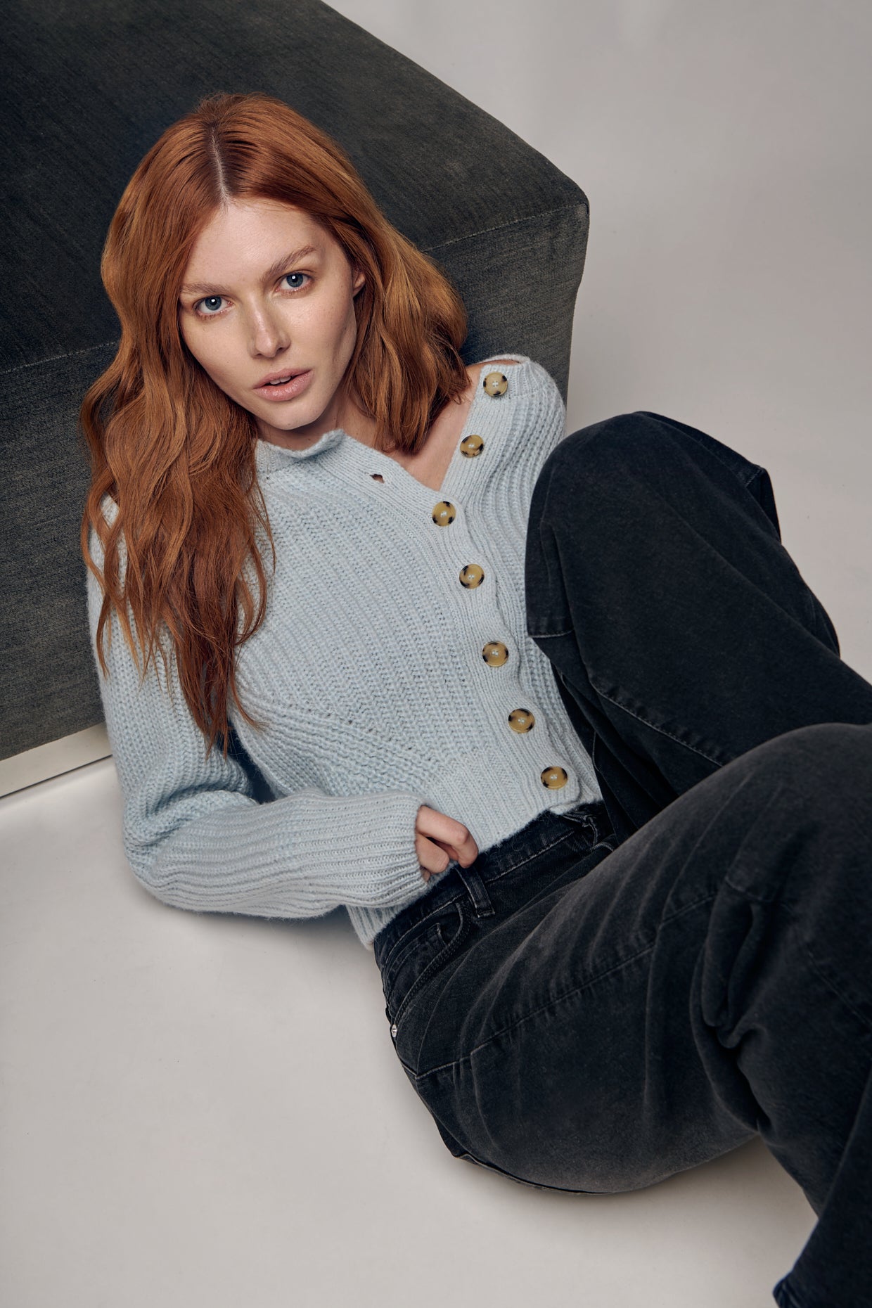 a woman leans against a sofa, exuding casual elegance in a chunky blue crew-neck cardigan from Eleven Six. The cardigan's soft blue hue and textured knit fabric are highlighted, adding depth to the ensemble. Paired with relaxed-fit denim, the look is effortlessly stylish and comfortable.