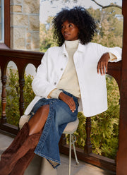 A woman with curly black hair sits on a stool on a stone balcony, wearing a white button-up jacket over a cream ribbed turtleneck sweater, blue jeans, and brown suede boots.