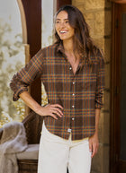 A woman wearing a brown plaid button-down shirt with orange and yellow stripes, paired with white pants, standing in a warm-toned stone doorway.