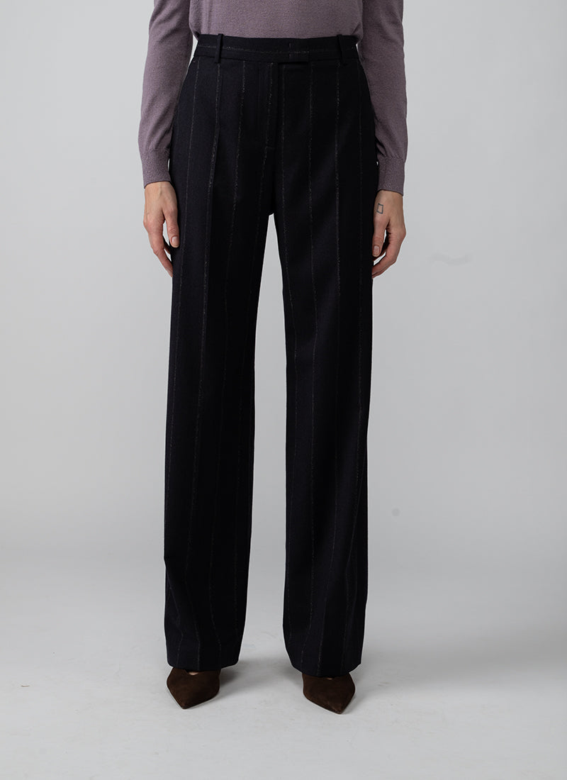 A model is shown from the waist down wearing high-waisted, wide-leg trousers in a very dark navy blue fabric with subtle vertical pinstripes. The model is also wearing a muted purple long-sleeve top and brown pointed-toe heels, standing against a neutral light gray studio background.