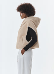 A model shown from the side profile wearing a beige fuzzy hooded vest over a black long-sleeve top and white pants, against a plain light gray studio background.