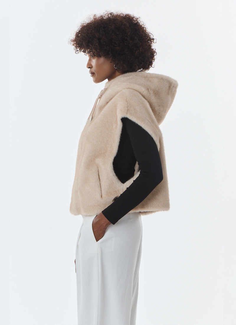 A model shown from the side profile wearing a beige fuzzy hooded vest over a black long-sleeve top and white pants, against a plain light gray studio background.