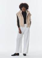 A model stands against a plain white studio background wearing a beige sherpa zip-up vest over a black long-sleeve top and white wide-leg trousers.