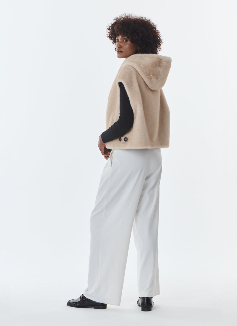 A model is shown from a side-back angle wearing a beige hooded shearling-style vest over a black long-sleeve top and white wide-leg trousers, against a plain white studio background.