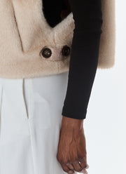 Close-up of a person wearing a beige fuzzy cropped jacket with dark buttons over a black long-sleeve top and white trousers, against a plain light background.