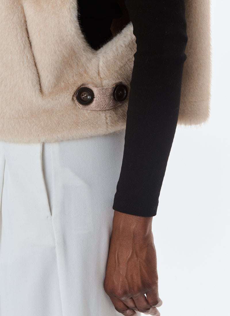 Close-up of a person wearing a beige fuzzy cropped jacket with dark buttons over a black long-sleeve top and white trousers, against a plain light background.