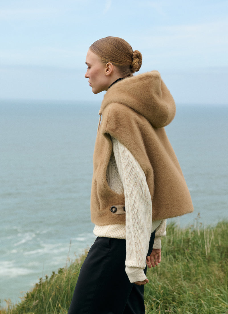 A woman stands in profile on a grassy cliff overlooking the ocean, wearing a camel-brown hooded sleeveless vest over a cream knit sweater and black pants.