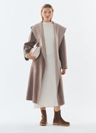 A woman modeling a long taupe-brown wide lapel coat over a cream dress, holding a matching taupe clutch, and wearing brown ankle boots against a plain light gray studio background.
