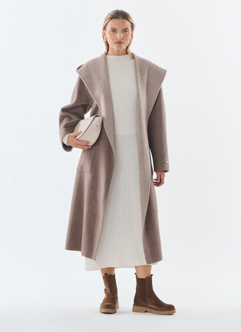 A woman modeling a long taupe-brown wide lapel coat over a cream dress, holding a matching taupe clutch, and wearing brown ankle boots against a plain light gray studio background.