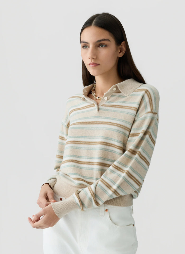 A woman modeling a beige and white striped knit polo sweater with thin brown and light blue horizontal stripes, paired with white pants, against a light gray studio background.