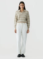 A model stands against a light gray studio background wearing a beige and white striped long-sleeve polo sweater, white wide-leg trousers, and black shoes.