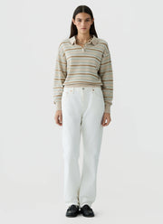 A model stands against a light gray studio background wearing a beige and white striped long-sleeve polo sweater, white wide-leg trousers, and black shoes.