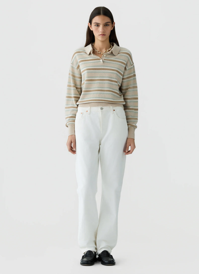A model stands against a light gray studio background wearing a beige and white striped long-sleeve polo sweater, white wide-leg trousers, and black shoes.