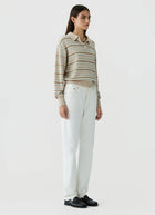 A woman stands against a plain light gray studio background wearing a beige and white horizontally striped long-sleeve polo shirt, white trousers, and black loafers.