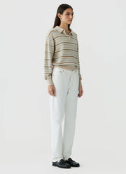 A woman stands against a plain light gray studio background wearing a beige and white horizontally striped long-sleeve polo shirt, white trousers, and black loafers.