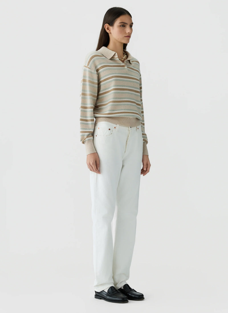 A woman stands against a plain light gray studio background wearing a beige and white horizontally striped long-sleeve polo shirt, white trousers, and black loafers.