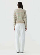 A woman viewed from behind wearing a beige and white striped knit sweater and white wide-leg trousers, standing against a light gray studio background.