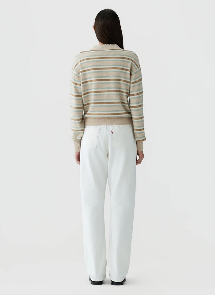 A woman viewed from behind wearing a beige and white striped knit sweater and white wide-leg trousers, standing against a light gray studio background.