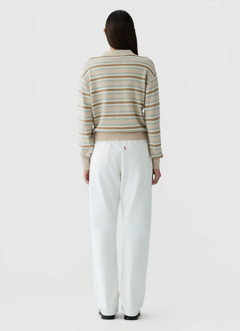 A woman viewed from behind wearing a beige and white striped knit sweater and white wide-leg trousers, standing against a light gray studio background.