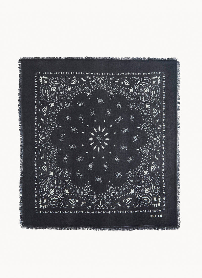 A square black bandana with white printed paisley and floral motifs, finished with fringed edges, shown flat against a light gray background. The brand name 'KUXTEN' is printed in small white letters in the bottom right corner.