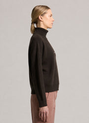 A woman in profile wearing a dark brown ribbed turtleneck sweater and dusty rose trousers, photographed against a neutral light gray studio background.