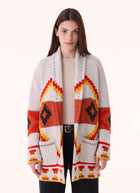 A woman modeling a long open-front knit cardigan with a Southwestern geometric pattern in orange, red, yellow, black, and off-white, worn over a light beige top and black pants against a plain light background.