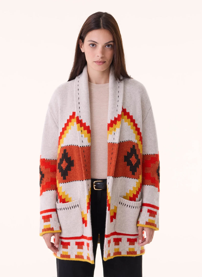A woman modeling a long open-front knit cardigan with a Southwestern geometric pattern in orange, red, yellow, black, and off-white, worn over a light beige top and black pants against a plain light background.