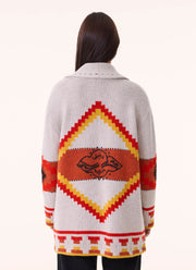 A woman viewed from the back wearing a cream knit cardigan with a large Southwestern-style geometric diamond pattern in red, orange, and yellow, and a black graphic motif in the center.