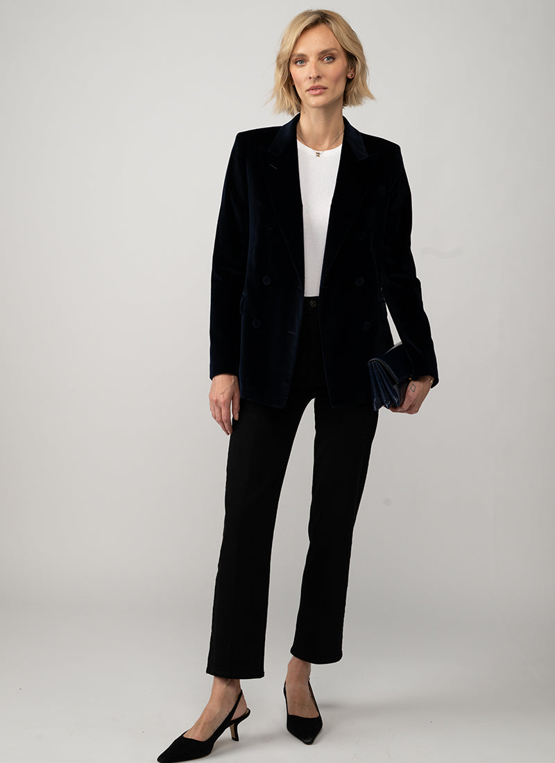 A woman modeling a black double-breasted velvet blazer with matching black trousers, a white top, and black heels, holding a small black velvet clutch against a light gray studio background.