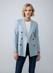 A woman modeling a light blue double-breasted blazer over a white top and dark blue jeans, photographed against a neutral light gray studio background.