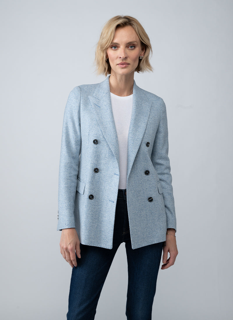 A woman modeling a light blue double-breasted blazer over a white top and dark blue jeans, photographed against a neutral light gray studio background.