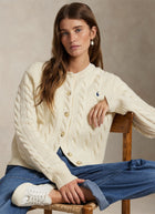 A woman seated on a wooden chair wearing a cream cable-knit cardigan with gold-tone buttons and a small embroidered polo player logo on the chest, paired with blue jeans and white sneakers against a neutral studio backdrop.